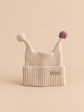 Gorro infantil de dos puntas con pequeños pompones, color beige, vista frontal en la fotografía del producto